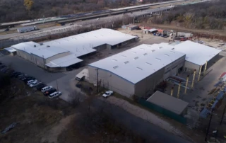Aerial view of a large industrial warehouse complex with bright white roofs, parking lots with cars, and a nearby highway bordered by trees.