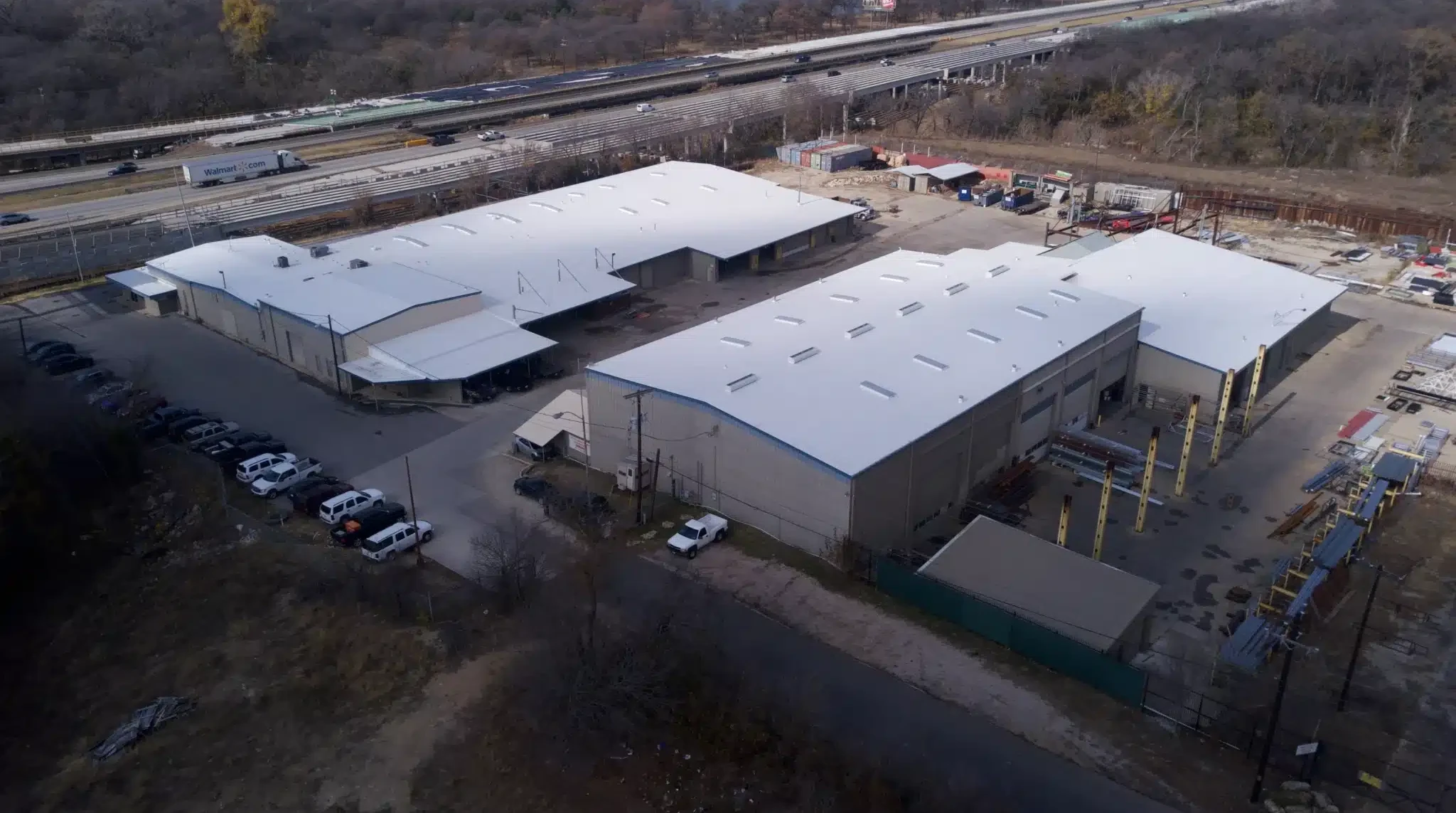 Aerial view of a large industrial warehouse complex with bright white roofs, parking lots with cars, and a nearby highway bordered by trees.