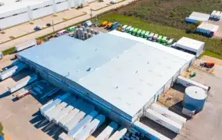 Aerial view of a large commercial roofing facility with loading docks, trailers, and rooftop drainage features.