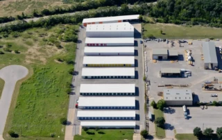 Aerial view of an industrial property with rows of white-roofed storage buildings and adjacent warehouses, bordered by trees and roads.