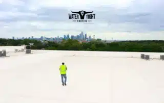 WaterTight Roofing technician walking across a white commercial roof during a roof coating inspection with a city skyline in the background.